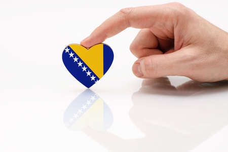Bosnia and Herzegovrina flag. Love and respect Bosnia and Herzegovina. A man's hand holds a heart in the shape of the flag on a white glass surface. The concept of Bosnian patriotism and pride.の写真素材
