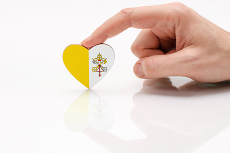 Vatican flag. Love and respect Vatican. A man's hand holds a heart in the shape of the Vatican flag on a white glass surface. Respect for Catholicsの写真素材