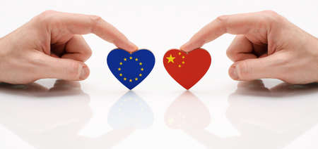 The concept of friendship and diplomatic relations between UE and the China. Two male hands are holding flags in the shape of a heart on a glass table. The flag of the European Union and the Republic of China as a symbol of cooperation and respect.の写真素材