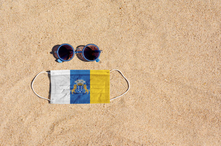 A medical mask in the color of the Canary Islands flag lies on the sandy beach next to the glasses. The concept of organizing a safe beach holiday in Canary Islands during the  pandemic.の写真素材