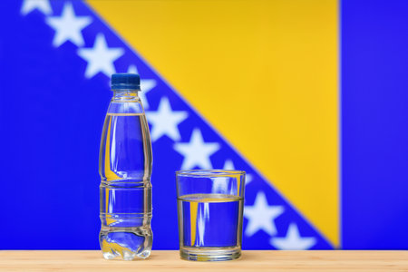 A bottle of clean drinking water and a glass stand on the table against the background of the flag of Bosnia and Herzegovina. Concept of clean drinking water supplies in Bosnia and Herzegovina.の写真素材