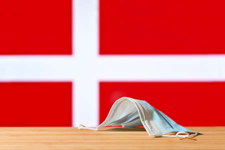 A medical mask lies on the table against the background of the flag of Denmark. The concept of a mandatory mask regime for residents of the country and tourists in Denmark during a pandemic.の写真素材