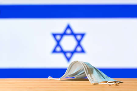 A medical mask lies on the table against the background of the flag of Israel. The concept of a mandatory mask regime for residents of the country and tourists in Israel during a pandemic.の写真素材