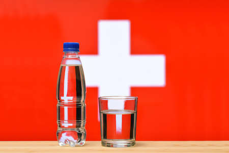 A bottle of clean drinking water and a glass stand on the table against the background of the flag of Switzerland. A concept for the supply of clean drinking water in Switzerland.の写真素材
