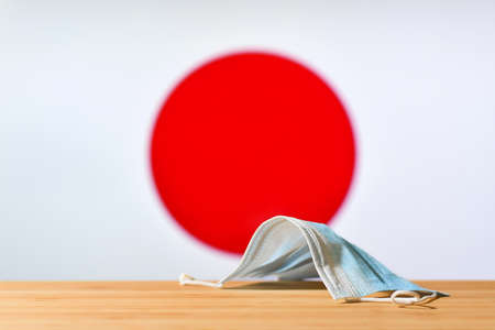 A medical mask lies on the table against the background of the flag of Japan. The concept of a mandatory mask regime for residents of the country and tourists in Japan during a pandemic.の写真素材