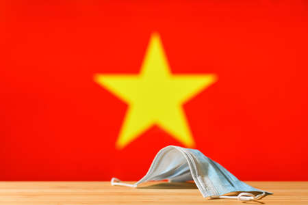 A medical mask lies on the table against the background of the flag of Vietnam. The concept of a mandatory mask regime for residents of the country and tourists in Vietnam during a pandemic.の写真素材