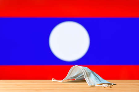 A medical mask lies on the table against the background of the flag of Laos. The concept of a mandatory mask regime for residents of the country and tourists in Laos during a pandemic.の写真素材
