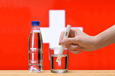 A hand with a water tester makes a measurement in a glass of clear water against the background of the flag of Switzerland. Test and assessment of drinking water supplies in Switzerland.の写真素材