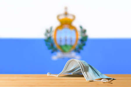 A medical mask lies on the table against the background of the flag of San Marino. The concept of a mandatory mask regime for residents of the country and tourists in San Marino during a pandemic.の写真素材