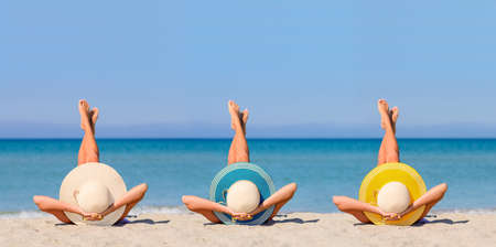 Three young girls on the beach wearing straw hats in the colors of the flag of Canary Islands. The concept of the perfect holiday in a Canary Islands resort. Focus on hats.の写真素材