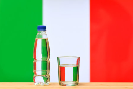 A bottle of clean drinking water and a glass stand on the table against the background of the flag of Italy. Concept of clean drinking water reserves in Italy.の写真素材