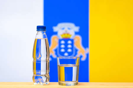 A bottle of clean drinking water and a glass stand on the table against the background of the flag of Canary Islands. A concept for the supply of clean drinking water in Canary Islands.の写真素材