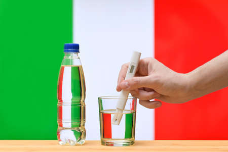 A hand with a water tester makes a measurement in a glass of clear water against the background of the flag of Italy. Test and assessment of drinking water supplies in Italy.の写真素材