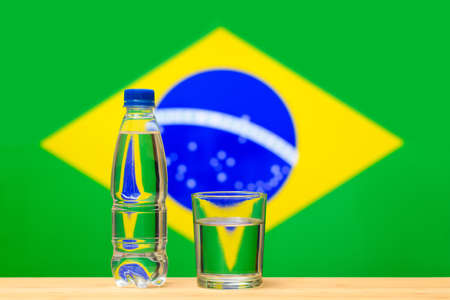 A bottle of clean drinking water and a glass stand on the table against the background of the flag of Brazil. A concept for the supply of clean drinking water in Brazil.の写真素材