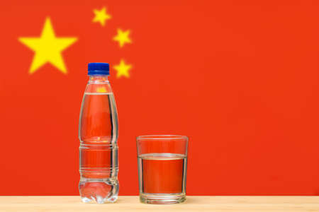 A bottle of clean drinking water and a glass stand on the table against the background of the flag of China. A concept for the supply of clean drinking water in China.の写真素材