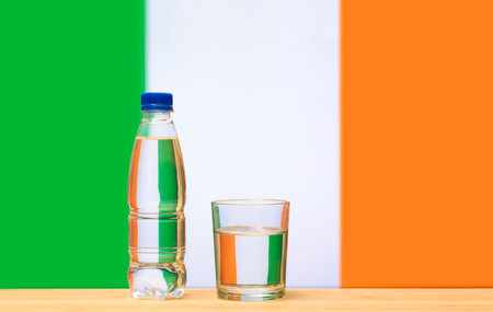 A bottle of clean drinking water and a glass stand on the table against the background of the flag of Ireland. A concept for the supply of clean drinking water in Ireland.の写真素材