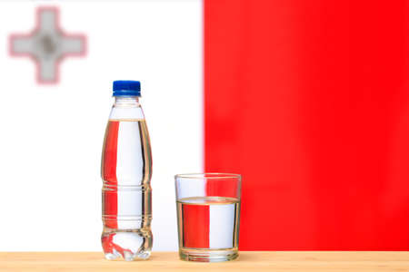 A bottle of clean drinking water and a glass stand on the table against the background of the flag of Malta. A concept for the supply of clean drinking water in Malta.の写真素材
