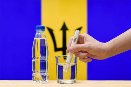 A hand with a water tester makes a measurement in a glass of clear water against the background of the flag of Barbados. Test and assessment of drinking water supplies in Barbados.の写真素材