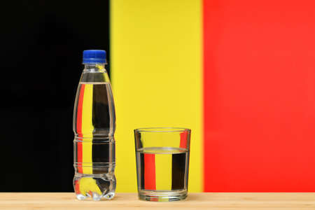 A bottle of clean drinking water and a glass stand on the table against the background of the flag of Belgium. A concept for the supply of clean drinking water in Belgium.の写真素材