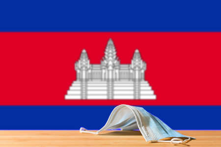 A medical mask lies on the table against the background of the flag of Cambodia. The concept of a mandatory mask regime for residents of the country and tourists in Cambodia during a pandemic.の写真素材