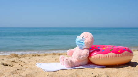 The concept of protecting tourists from the spread of coronavirus during a sea vacation. Soft toy in a mask on a sandy beach against the background of the sea.の写真素材