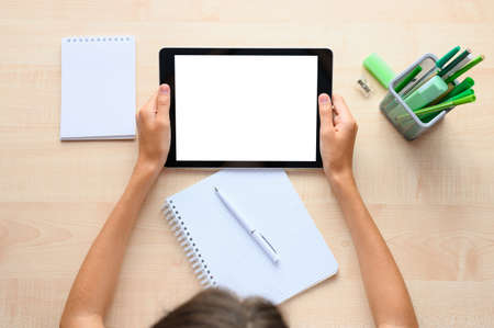 A girl at a school desk with a tablet in her hands. Tablet with blank white display. Top view,...