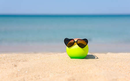 A colored ball with wearing heart-shaped sunglasses lies on a sandy beach against the backdrop of the sea.の写真素材