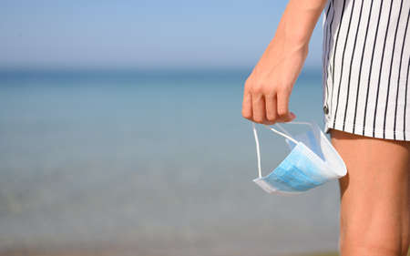 A young girl in a white dress stands on the beach and holds a medical mask in her hand.の写真素材