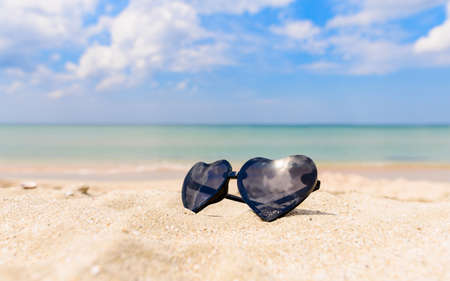 Sunglasses in the shape of a heart lie on a sandy beach on a sunny day against the backdrop of the sea. Sea travel concept. Selective focusing.の写真素材