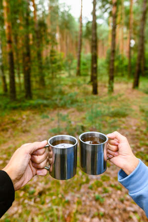 A guy and a girl on a picnic in the forest are holding mugs of tea. Vertical frame. Selective focus on the male handの写真素材