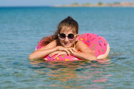 A cheerful little girl bathes in the sea on an inflatable pink circle. Portrait of a joyful girl on vacationの写真素材