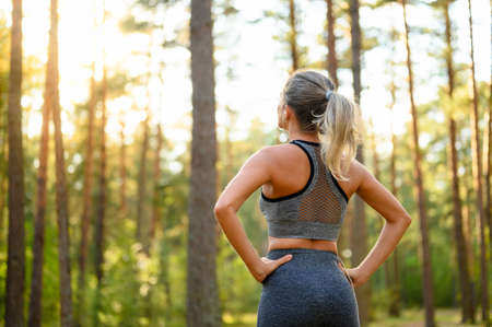 A sportive girl stands in the forest before a fitness class and looks towards the sun. View from the back. Selective focusing.の写真素材