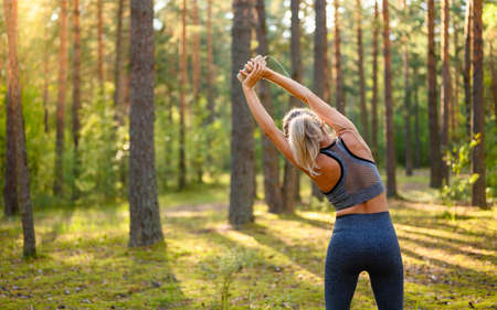 The girl makes bends to the side in the fresh air. The girl performs a warm-up before jogging. View from the back.の写真素材