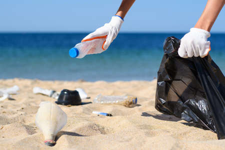 Close-up girl collects plastic garbage on the beach and puts it in a bag. Selective focus on the hand with the bottle.の写真素材
