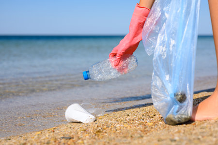 The girl collects plastic trash floating in the sea. Plastic debris floated to the shore. Selective focus on the hand with the bottle.の写真素材
