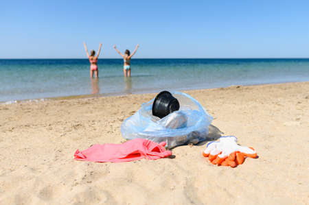 A full garbage bag and protective gloves lie on a clean beach. In the background, happy girls swim in the clear sea after cleaning up the garbage. Selective focus on debris.の写真素材