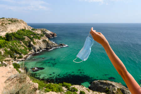 Medical mask in the outstretched hand of a girl against the backdrop of a beautiful seascape. Healthy vacation concept. Selective focus on handの写真素材