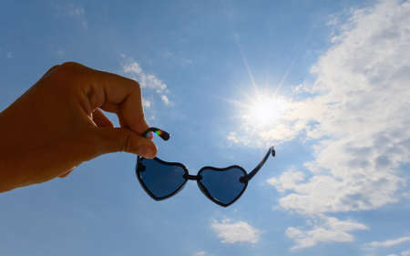 Sunglasses in the shape of hearts in a hand against a background of sky and sunbeams. Selective focusing on glasses.の写真素材