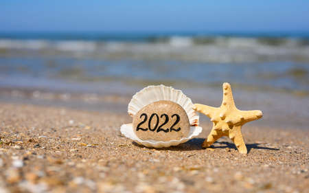 A stone with the inscription of the new year 2022 stands inside a large white shell next to a starfish. In the background the sea. Selective focus on the stone.の写真素材