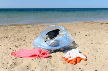 A full bag of plastic waste collected from the beach and protective gloves on the sand. Selective focusing.の写真素材