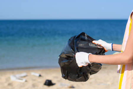 Garbage bag in hands against the background of a dirty seashore. The concept of preparing for the collection of garbage on the beach. Selective focusing.の写真素材