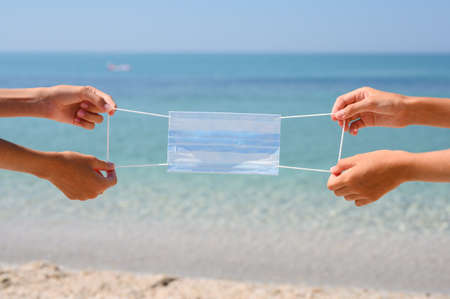 Medical mask in the hands of two tourists against the background of the sea. Vacation coronavirus protection concept.の写真素材