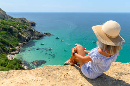 A young girl in a straw hat sits on the edge of a cliff and looks at the beautiful seascape with yachts and a transparent sea. Travel across the Crimea.の写真素材
