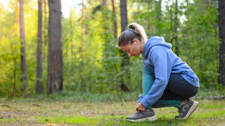 A girl in the woods prepares for her morning run and ties her shoelaces. Side view. Free space for text on the right.の写真素材