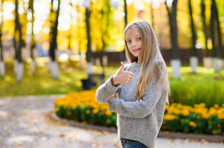Portrait of a joyful girl with long hair and in a knitted sweater against the background of an autumn park. The girl shows a thumbs up gesture.の写真素材