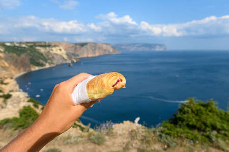 Sweet roll with jam in a man's hand against the backdrop of a beautiful seascape. Traveler's picnic on the background of the mountains and the sea.の写真素材