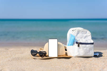 A smartphone with a blank white display on a sandy beach next to a backpack with a medical mask and a straw hat. In the background the sea.の写真素材