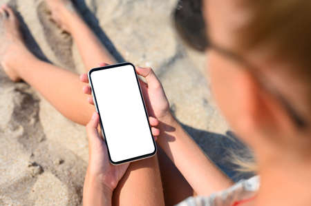 A girl sits on the beach and holds a smartphone with a blank white display in her hands. View from above.の写真素材