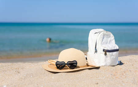Sunglasses in the shape of hearts, a straw hat and a white female backpack on a sandy beach. In the background, a girl is swimming in the sea. Selective focus on objects on the beach.の写真素材