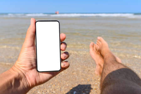 A man lies on the beach next to sea water and holds a smartphone with a blank white display in his hand. In the background, the sea and male legs.の写真素材